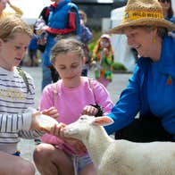 Three people crouched down with one holding a bottle feeding a lamb