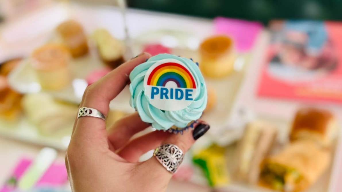 Pride Month - Afternoon Tea Trip - a hand holding out a small cup cake with swirl of icing on and a small rainbow logo, against blurry background of tea setting