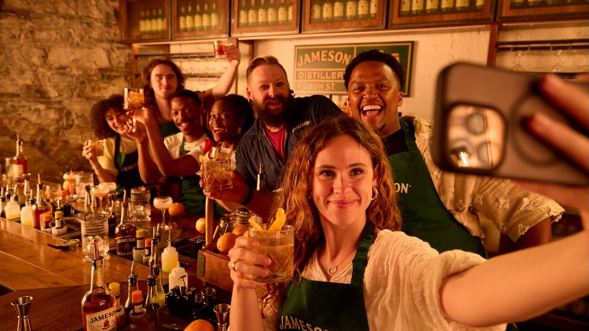 Group of smiling people at a bar raising a glass with woman at the front taking a group selfie.