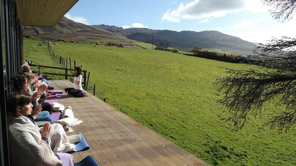 Group of people on sitting on yoga mats on a wooden decking facing a field
