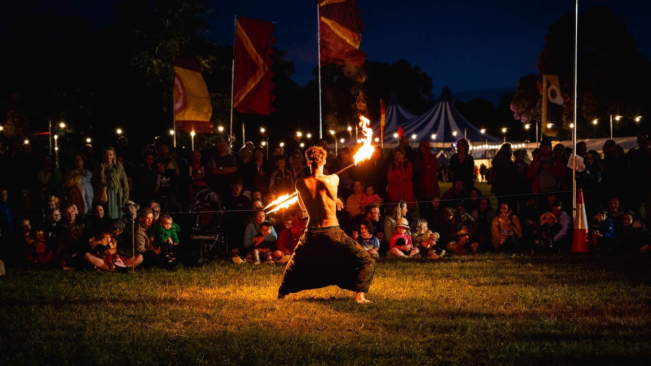 A night a crowd are watching a shirtless man who is moving with a fire stick.