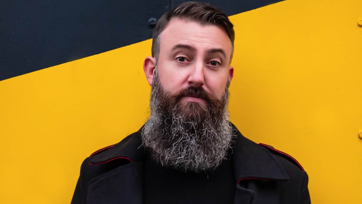 A man with long beard standing against bright yellow wall