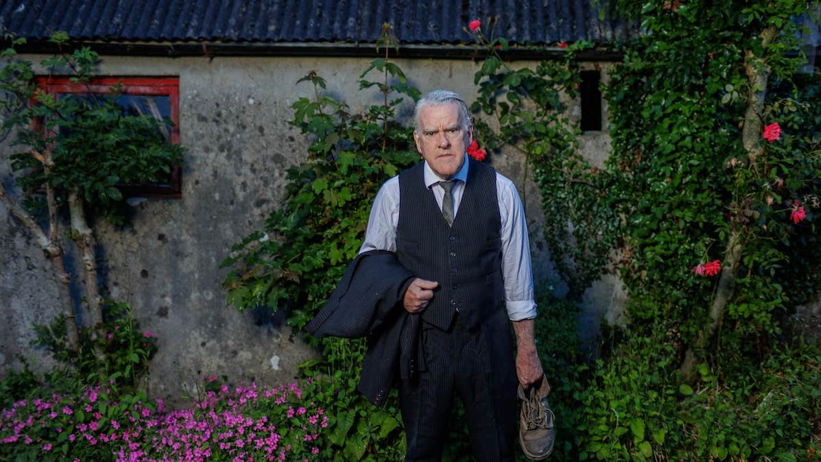 A cross looking man in shirt and tie is holding an old shoe and his jacket, standing in front of an old cottage