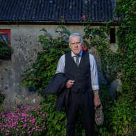 A cross looking man in shirt and tie is holding an old shoe and his jacket, standing in front of an old cottage