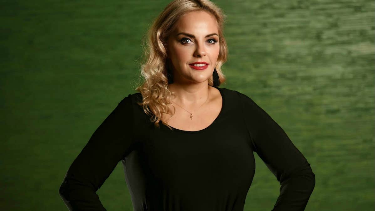 A gently smiling woman in black top with dark green background.