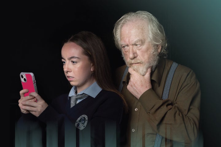 A teenager in school uniform is looking with concert at a mobile phone, a older man stood behind her looking over her shoulder at the phone is holding his hand up to his chin also looking serious.