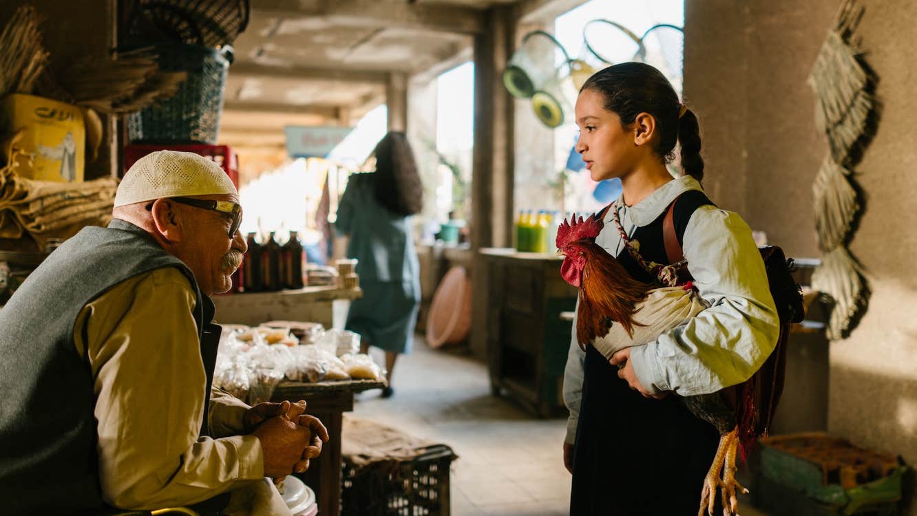 A girl with a chicken is talking a man at a market stall.