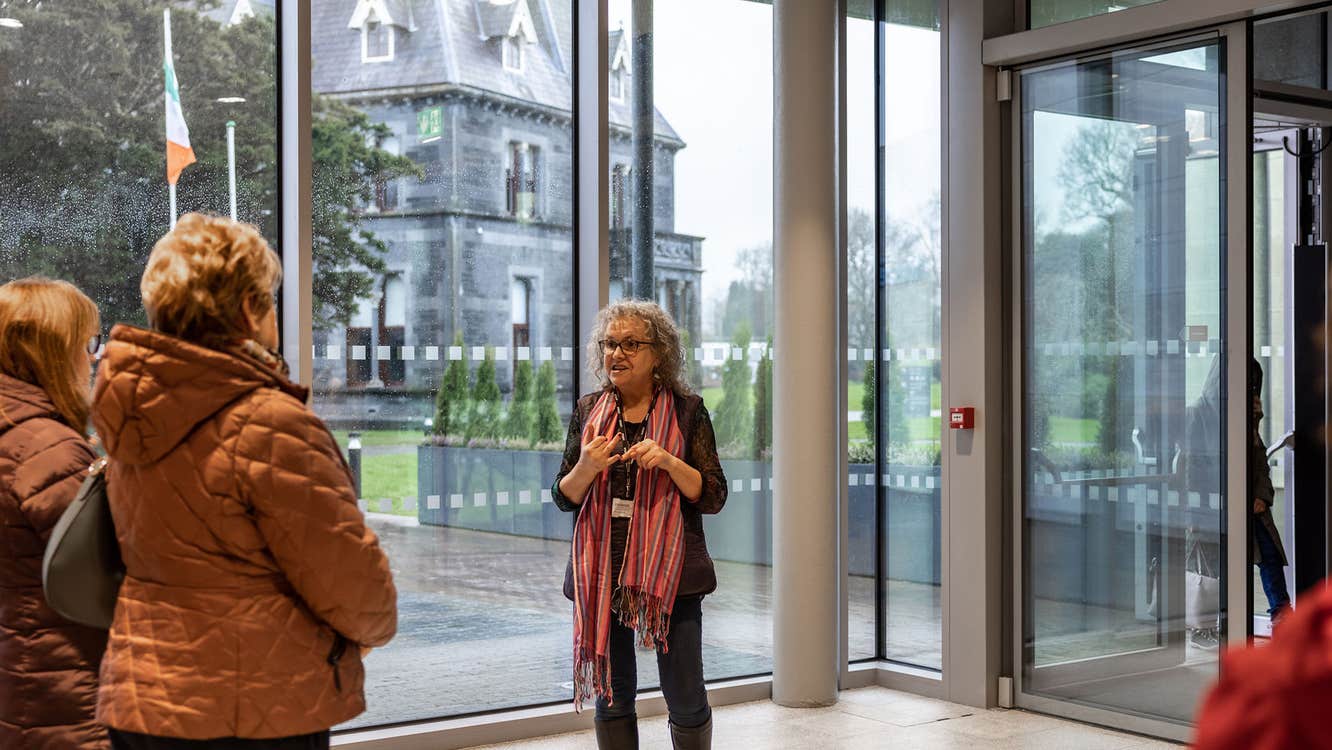 A woman tour guide in a glass lobby area is talking to small group with old, grey building visible through glass wall.