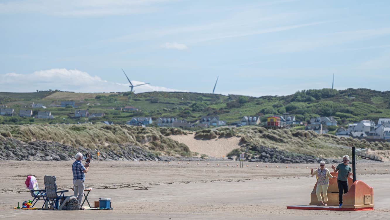 View of a sandy beach with 2 people dancing on small area, another person watching from a distance with deck chair.