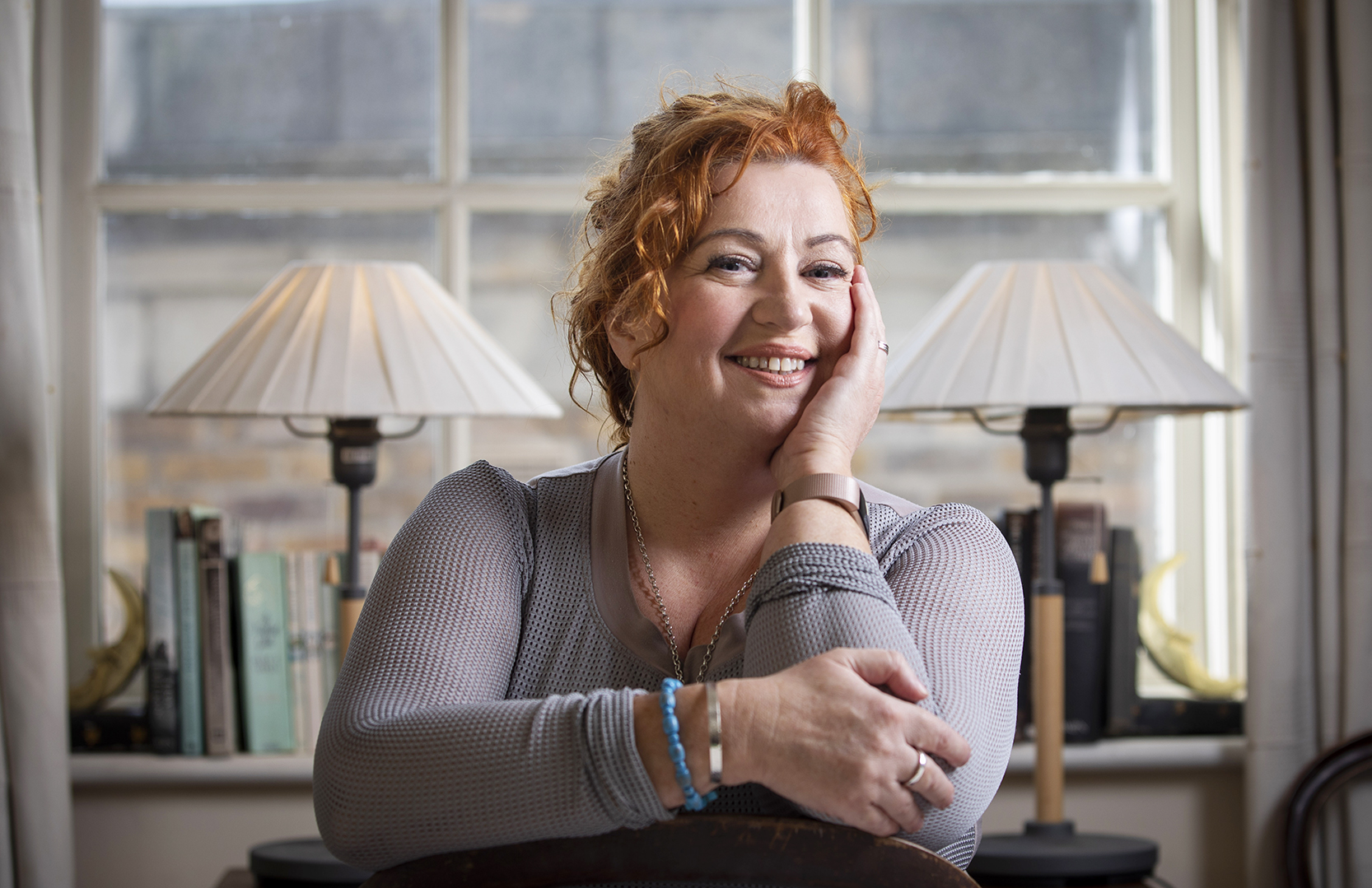 A smiling woman is resting face on left hand with arms crossed, against background of window with 2 table lamps either side in front of window and some books.