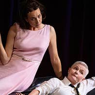 THE PATIENT GLORIA by Gina Moxley. A woman in a pink dress sitting on the arm of an armchair next to a women dressed in shirt and tie, on stage with a black background
