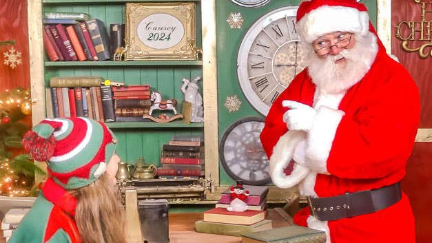 A santa is pointing a finger at a young elf seated at a wooden table with book shelf in background.