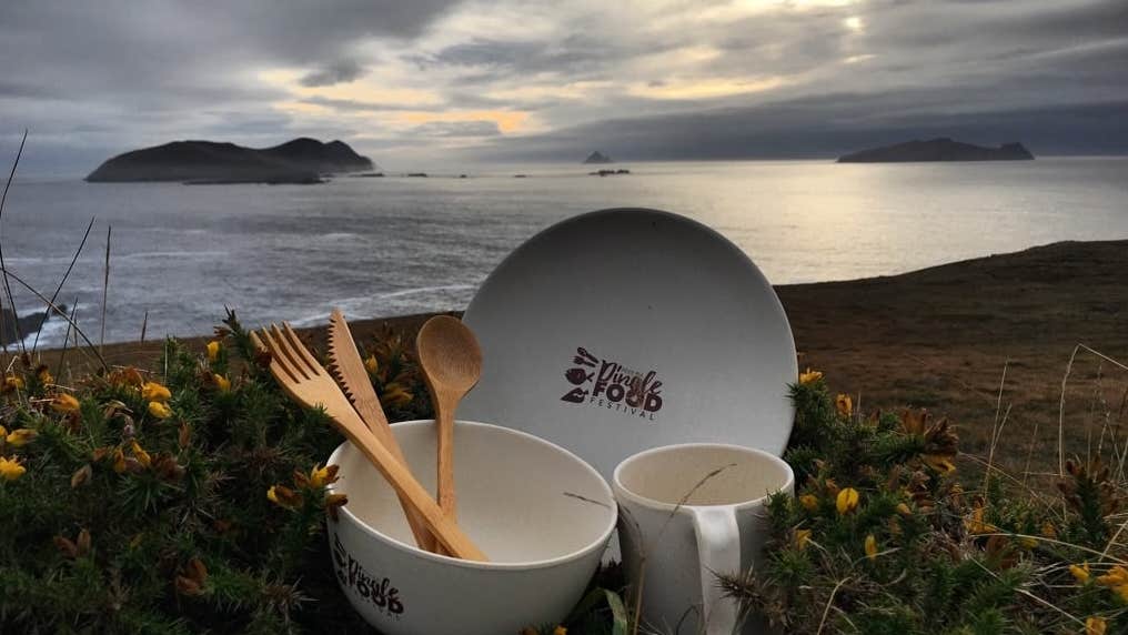 A white bowl, plate and mug holding set of wooden cutlery set against sea and rocky background.
