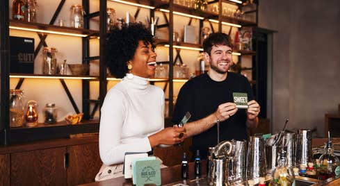 A woman and a man laughing as they show cue cards to people across a bar from them on a whiskey tour.