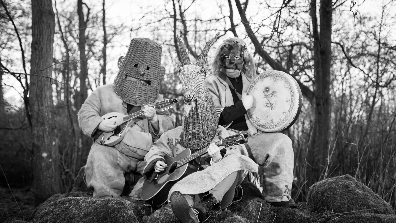 Black and white photo of 3 people sitting on rocks against background of bare trees, all have large masks covering their faces, holding musical instruments.