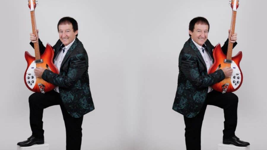 A smiling man in dark suit with one foot up on small white block, holding up a guitar vertically