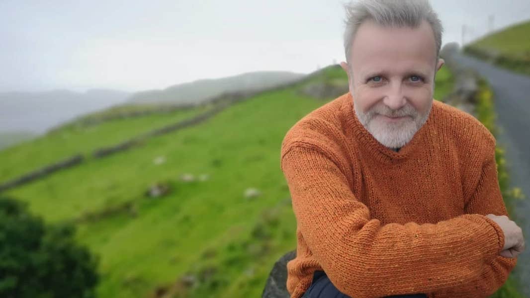 A smiling man with crossed arms outdoors with background of green hills