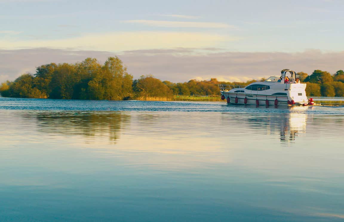 A boat sailing down a calm river near the shore on the Shannon