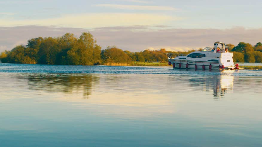 A boat sailing down a calm river near the shore on the Shannon