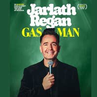 Man dressed in black, smiling holding a mic up to his face, against dark green background.