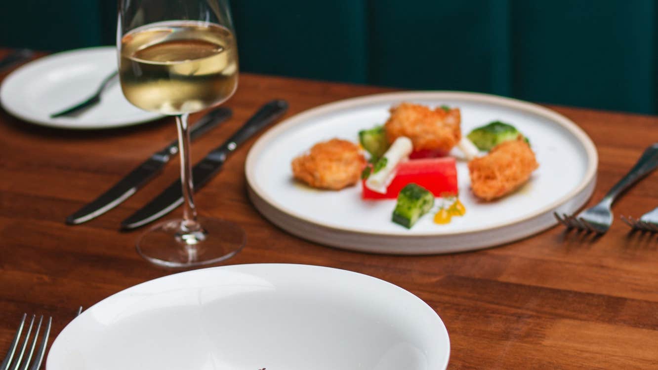 Table with two white plates of food a glass of wine and cutlery on it