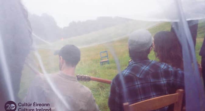 rear view of 3 people seated on wooden chairs in a grassy field, viewed through white net curtain.