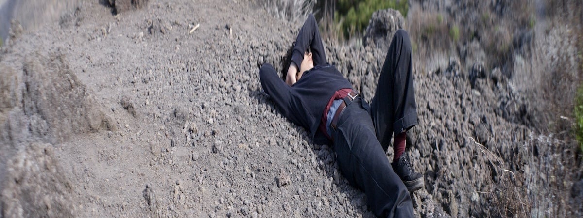 still of a woman dressed in all black lying on the slopes of Mount Vesuvius. Her right hand is behind her hand is behind her head and she raises her left arm over her head.