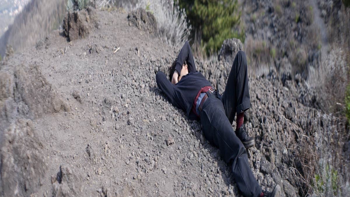 still of a woman dressed in all black lying on the slopes of Mount Vesuvius. Her right hand is behind her hand is behind her head and she raises her left arm over her head.