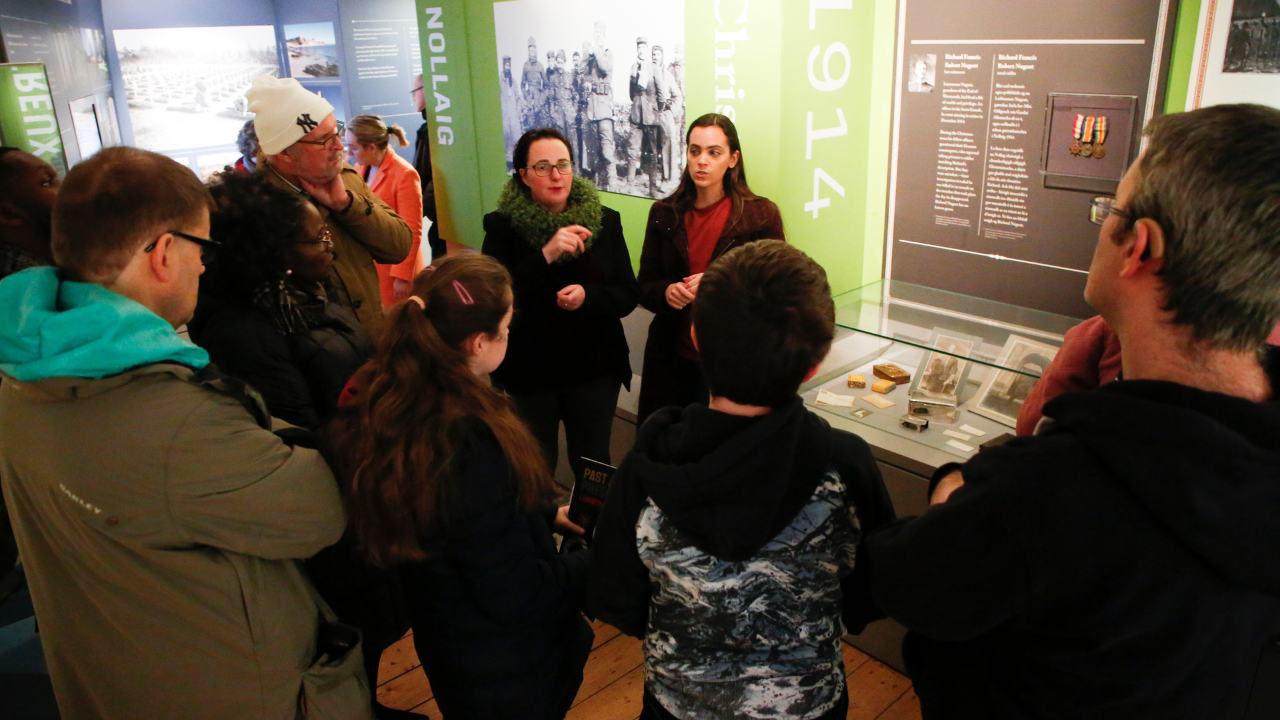 A small group of people are gathered around 2 women, standing in front of a small display cabinet in a museum.