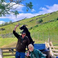 A couple smile at the camera as a goat playfully climbs behind them