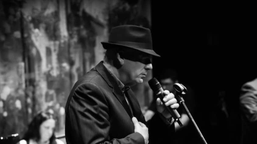 Black and white photo, side view of man in suit and hat singing into a mic.