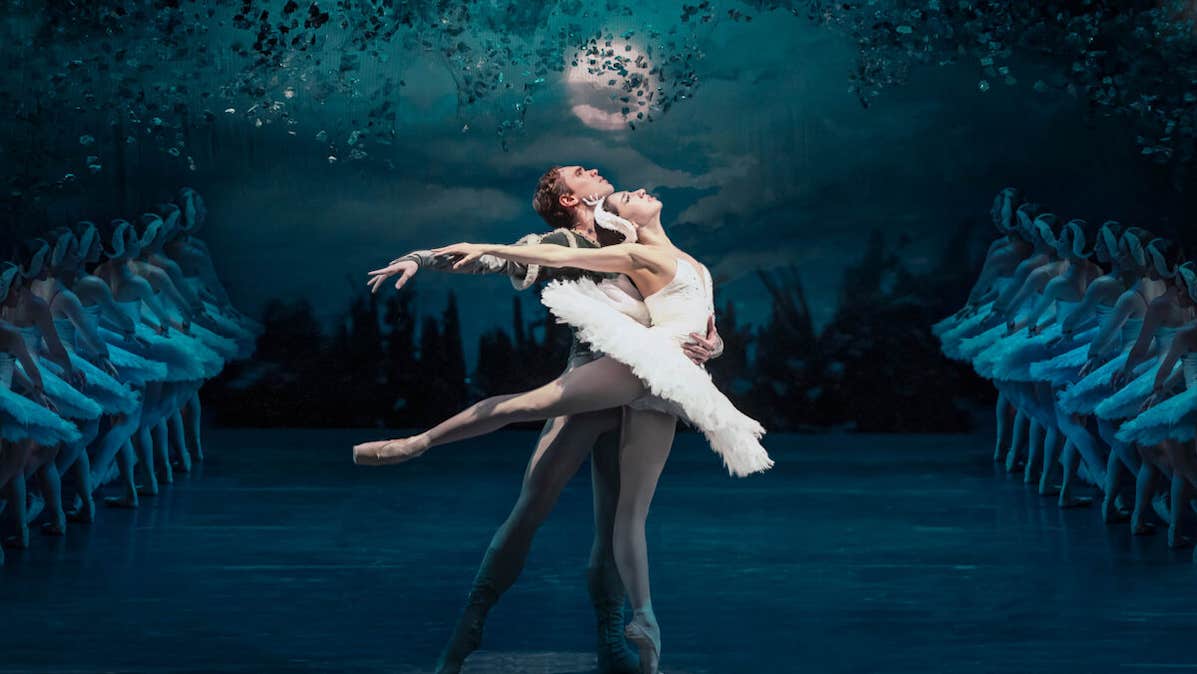 A female in short, white tutu on point ballet shows being supported by a man beside her on a dark stage