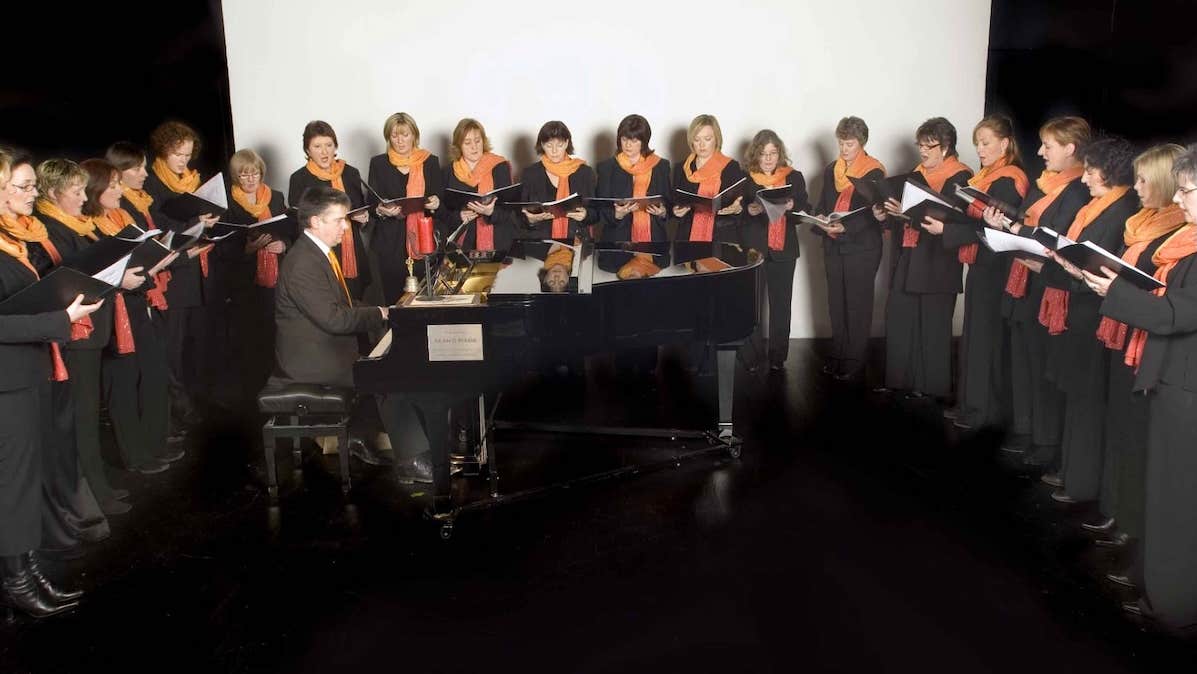 Festival of Voice: Cór Ban Cúil Aodha, a group of women standing in a semi circle around a small grand piano being played by a man.