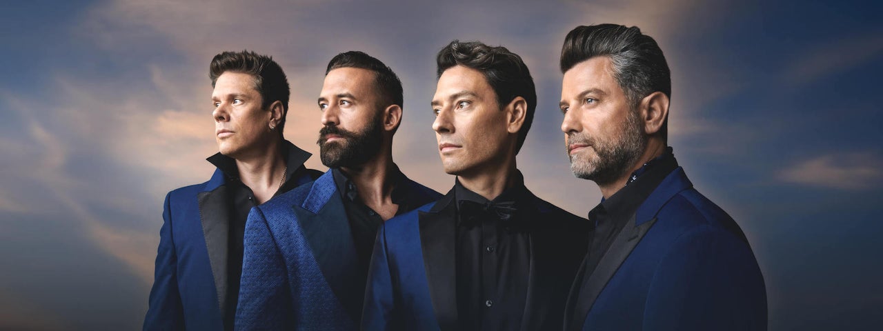 4 men in dark blue suits in a line all looking away to their right against murky sky.