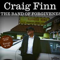 A man in jacket, white shirt and peaked cap is leaning against a silver car outside shops