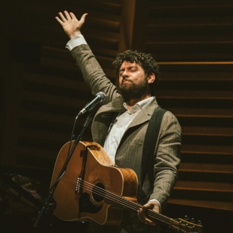 A man standing with a guitar has his eyes closed with his right arm up in the air.