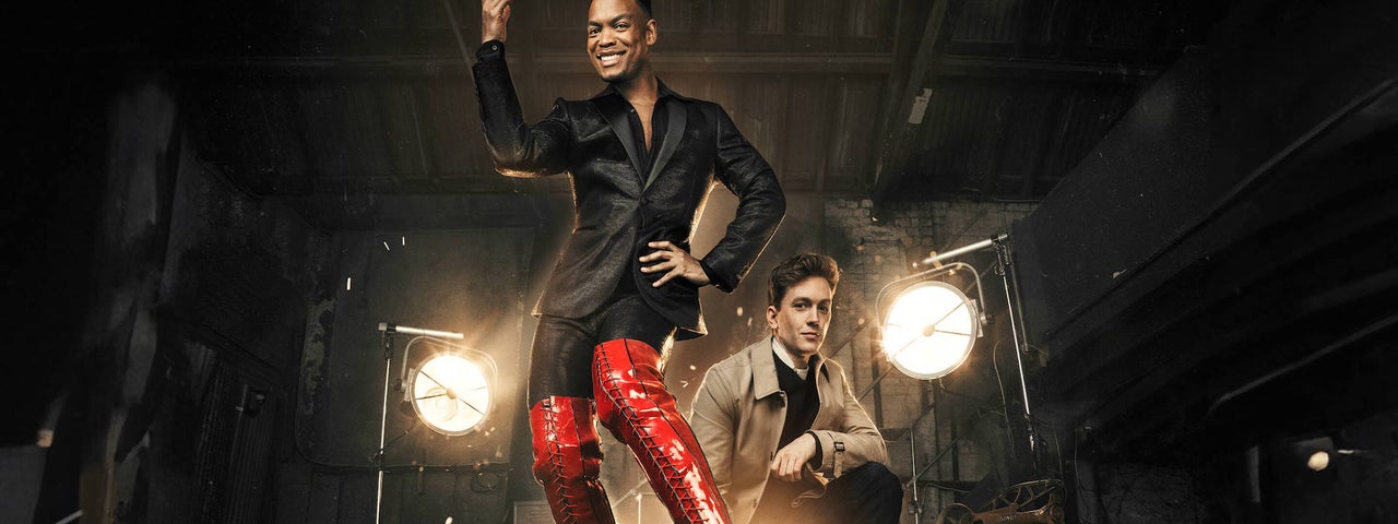 Kinky Boots The Musical, a smiling person in black wearing tall red shiny boots on a stage with another man crouching down behind him.
