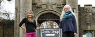 Two ladies standing outside a castle like gate lodge beside a sign that says crafts with an arrow pointing to the right