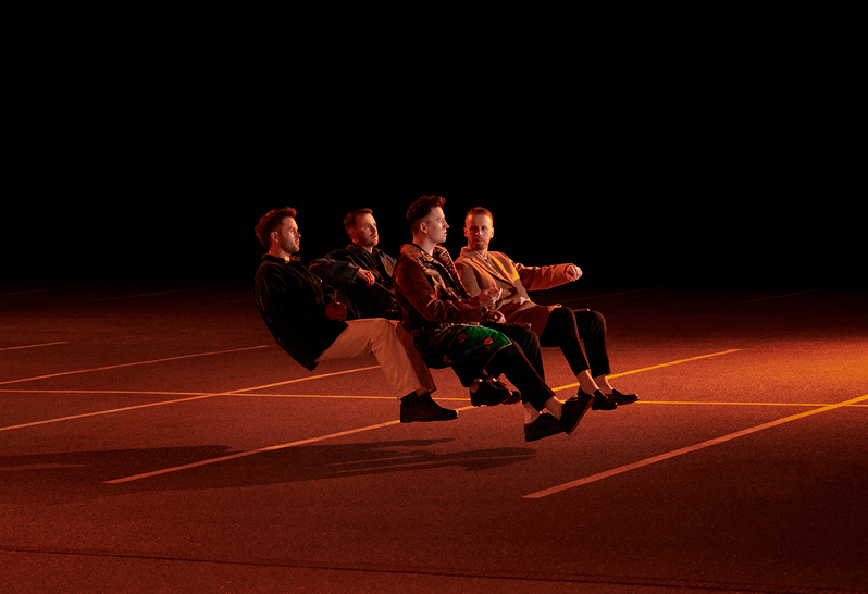 Picture This at the INEC Arena. Darkly lit photo of 4 people appearing to be sitting in a car that doesn't exist, hovering over empty car parking spaces.