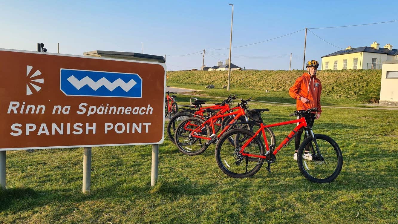 Child in an orange hoodie standing on grass by some red bikes and a road sign