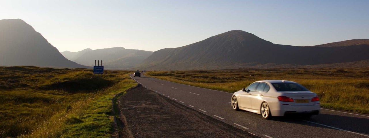 White car driving on a wide road towards mountains with green landscape on either side