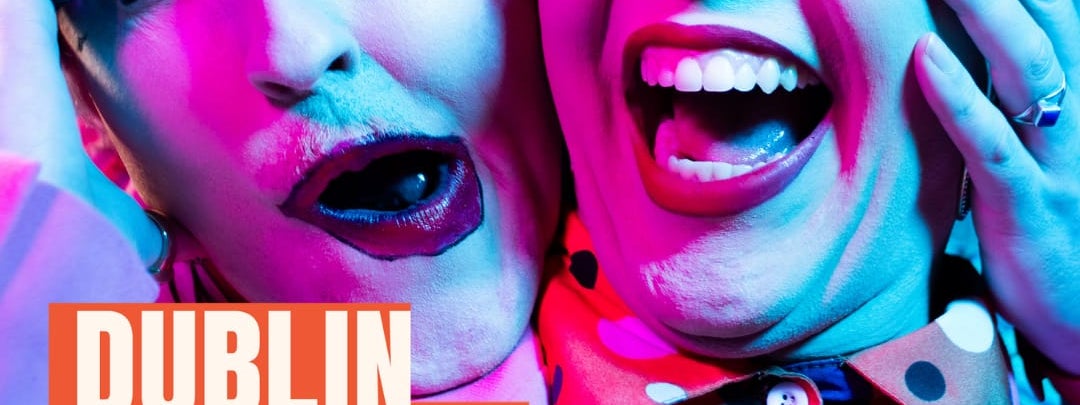 Two drag queens pose side-by-side, holding each other's faces and smiling into the camera. They wear sharp blonde wigs and colourful make-up. The queen on the right wears glasses. Dublin Fringe Festival's logo is in the bottom-left corner.