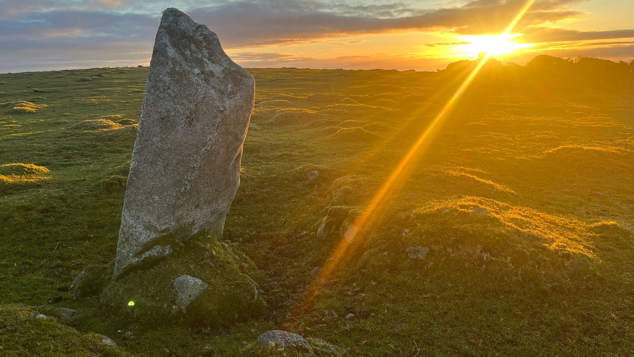 A granite standing stone, with a sunrise breaking through cloud behind it