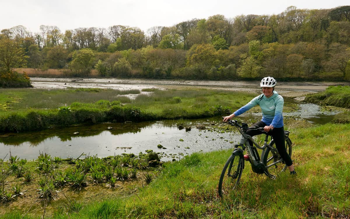 Person sitting on a bike in a field next to a river with trees in the background