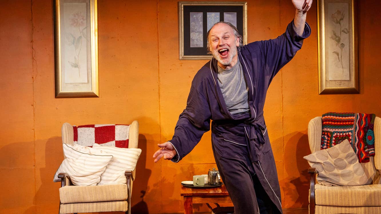 On a stage with armchairs, a man in a dressing gown is waving his arms looking excited.