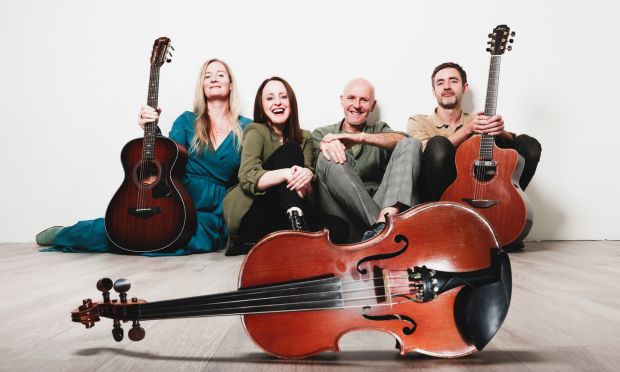 Music Network presents Niamh Dunne, Bróna McVittie, Cormac Breatnach & Seán Óg Graham live in concert. 4 people are sat on the floor against a plain wall, smiling and laughing with guitars and fiddle.