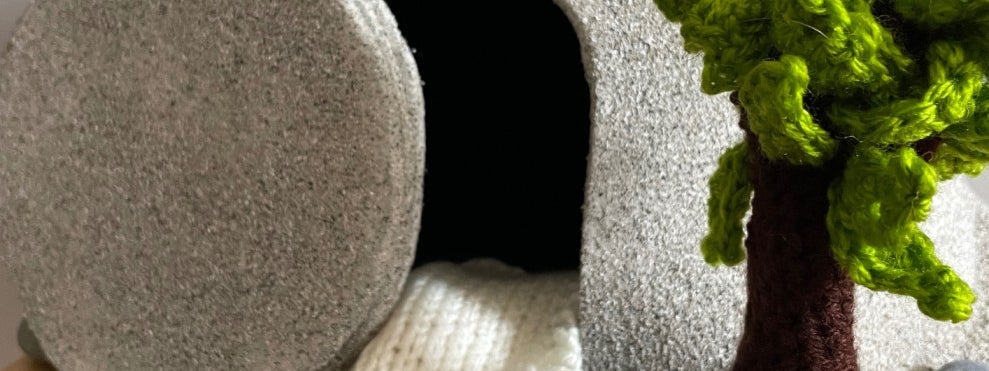 A stone tomb, open, with knitted trees and smaller stones surrounding it.