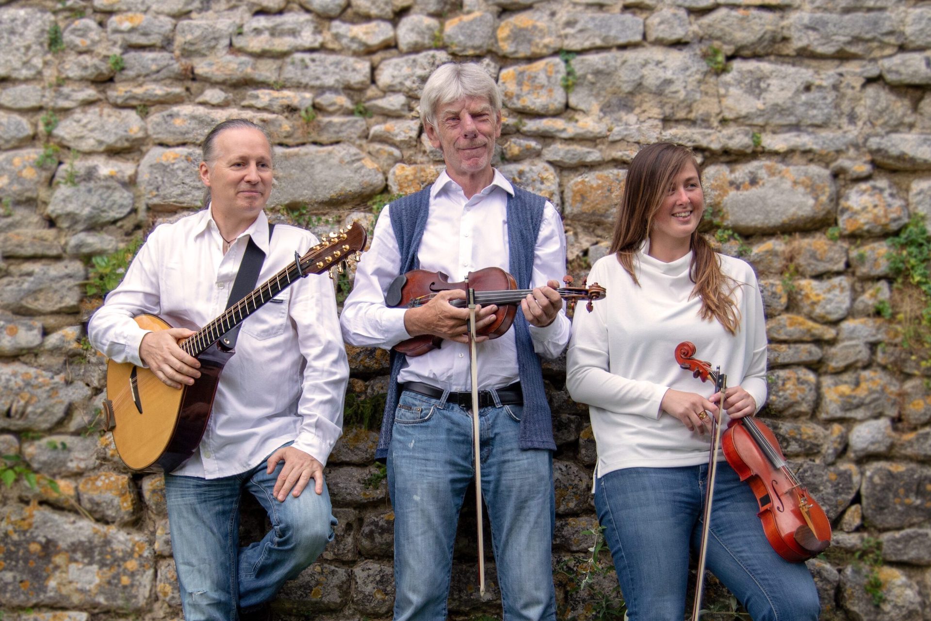 3 people smiling, leaning against an old stone wall holding musical instruments, dressed in white shirts and jeans.