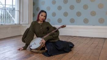 A woman with stringed instrument seated on a bare wooden floor in an empty room