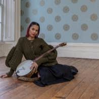 A woman with stringed instrument seated on a bare wooden floor in an empty room